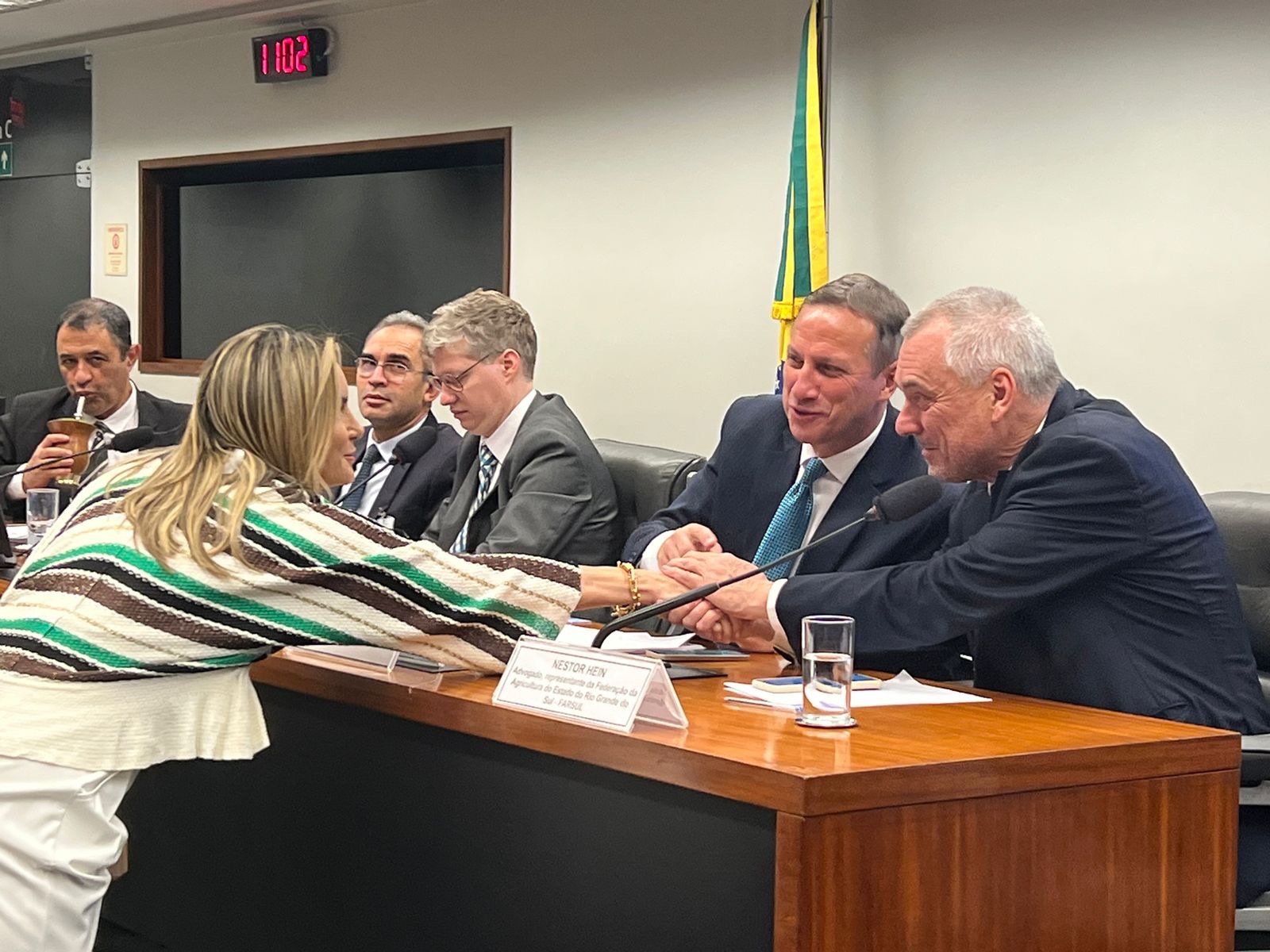 Encontro institucional em sala de reunião, com aperto de mãos sobre a mesa, ao lado de placas de identificação de representantes