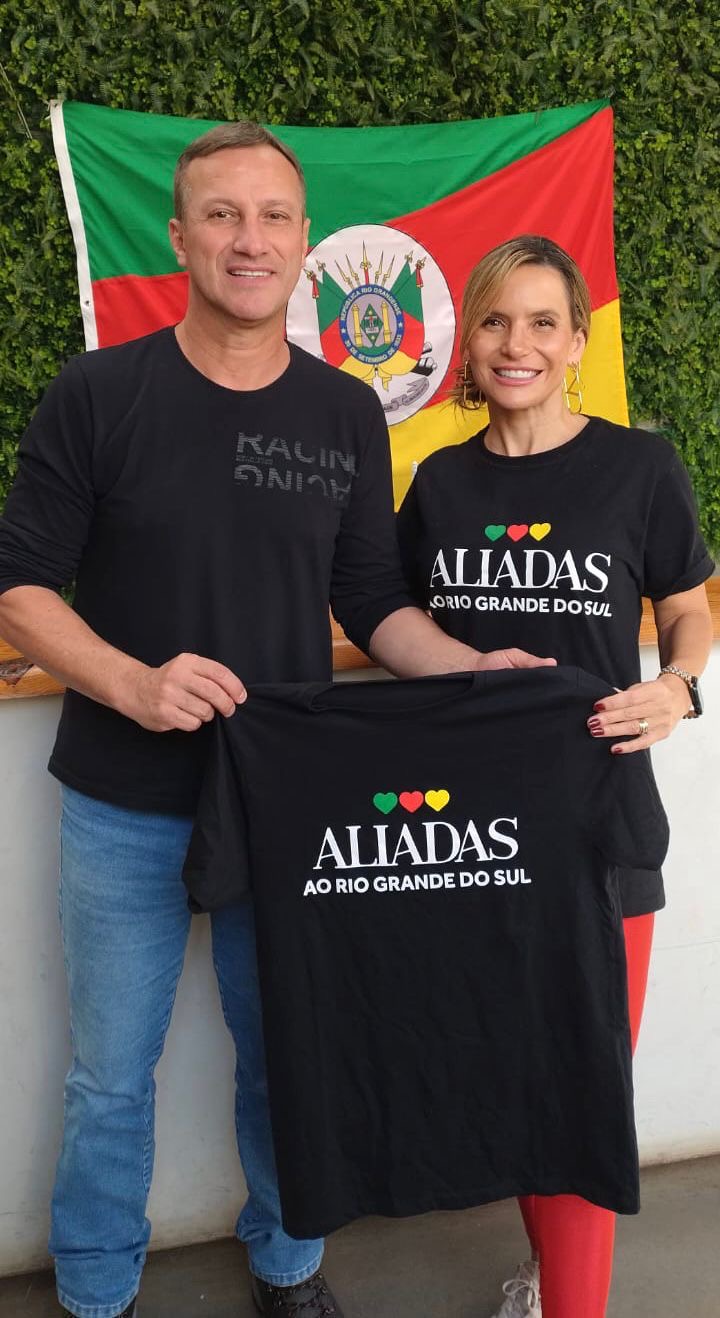 Homem e mulher seguram uma camiseta com a mensagem 'Aliadas ao Rio Grande do Sul', posados diante da bandeira do estado