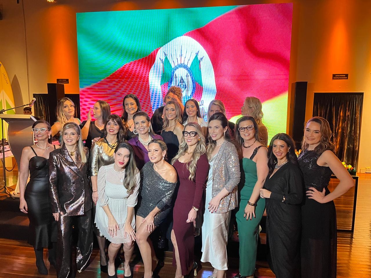 Foto coletiva de mulheres do Instituto Aliadas reunidas no evento, diante do painel com a bandeira do Rio Grande do Sul