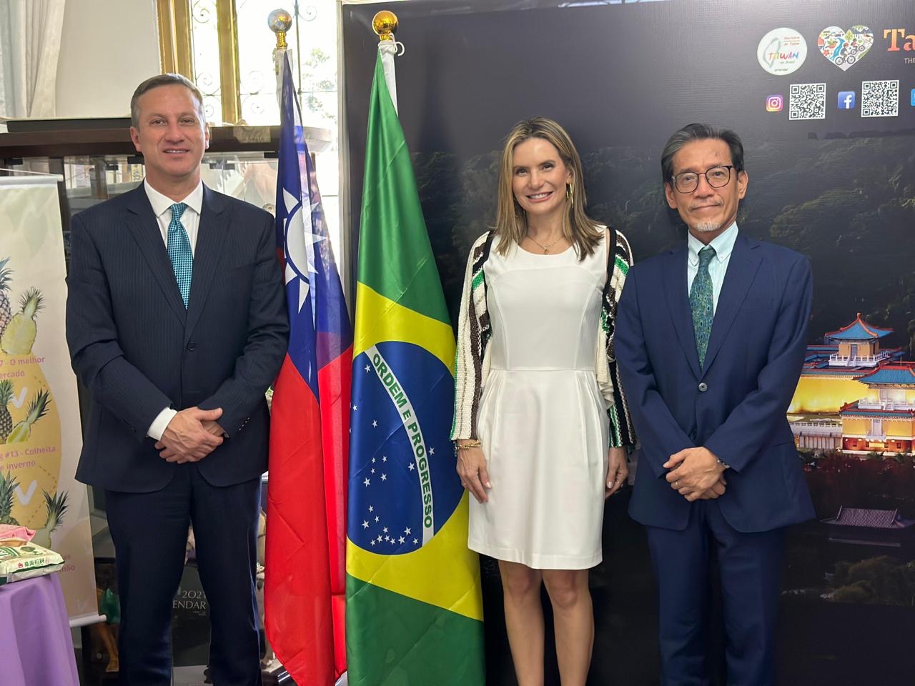 Três pessoas em traje formal posam para fotografia em ambiente oficial, entre as bandeiras de Taiwan e do Brasil, com painel de identificação do Governo de Taiwan ao fundo