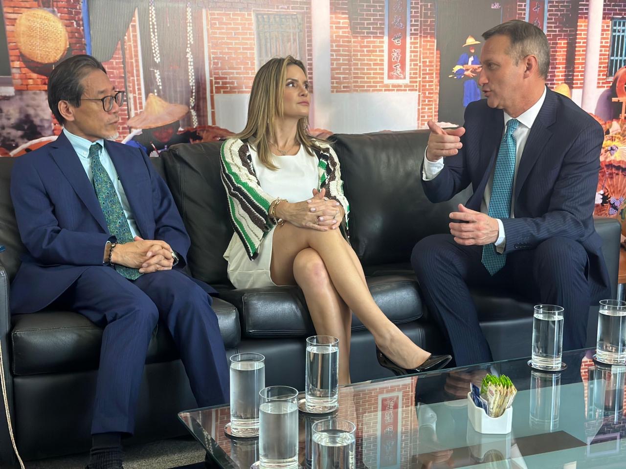 Três pessoas em traje formal conversam em ambiente oficial, sentadas em um sofá diante de um mural