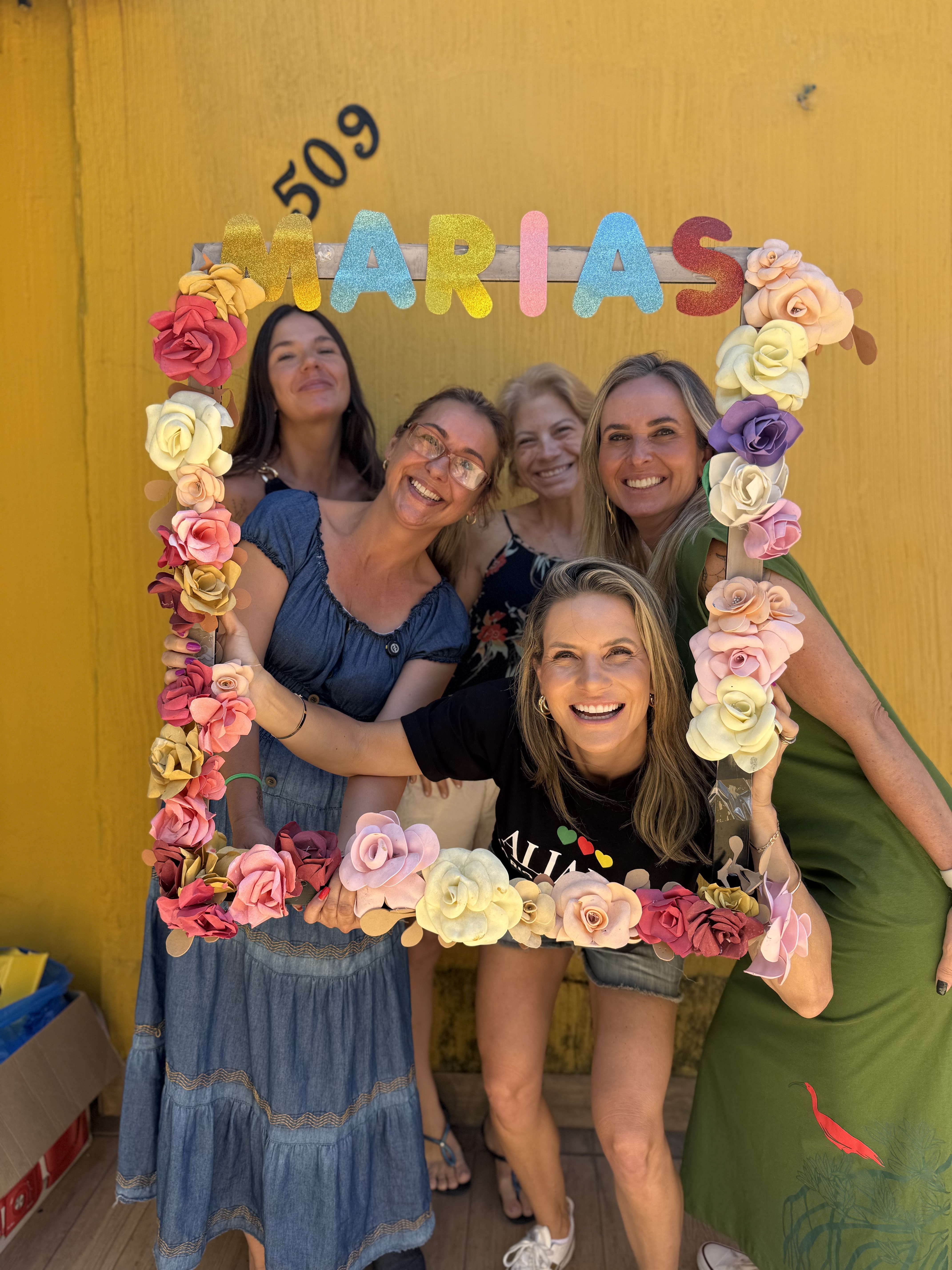 Cinco mulheres posam juntas segurando uma moldura artesanal com a palavra MARIAS, em frente à parede amarela da sede revitalizada do Grupo Marias