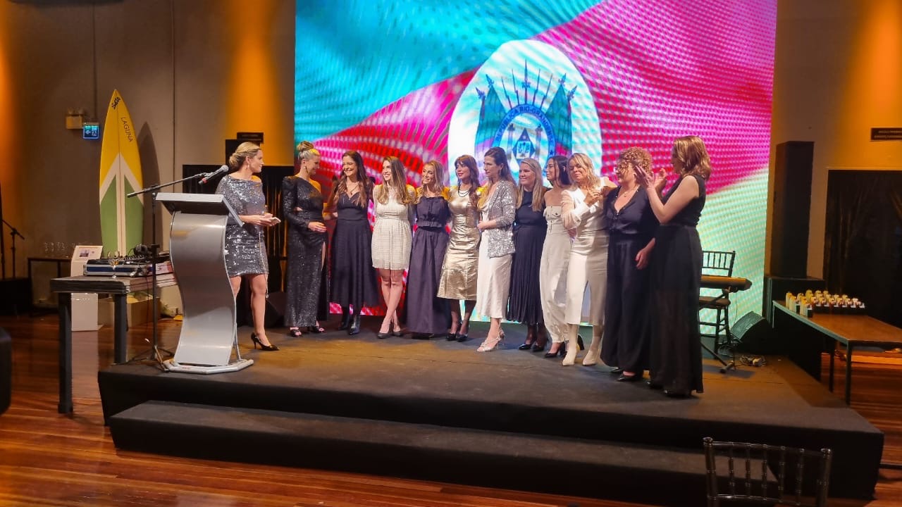 Grupo de mulheres reunidas no palco durante a cerimônia, ao lado de um púlpito, diante do painel com a bandeira do Rio Grande do Sul