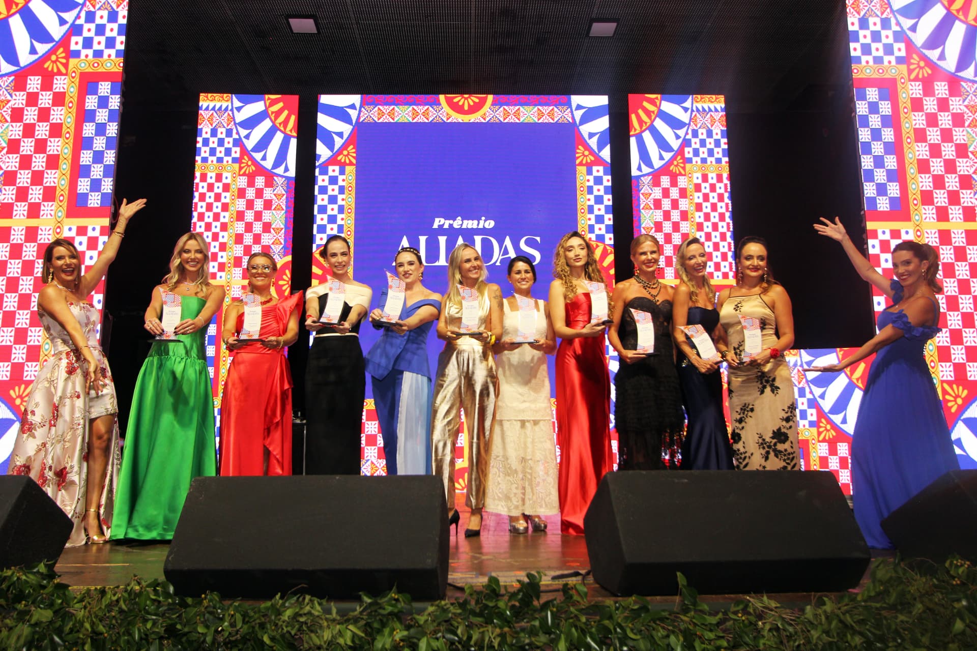 Homenageadas do Prêmio Aliadas 2025 reunidas no palco segurando seus troféus, com o painel do evento ao fundo