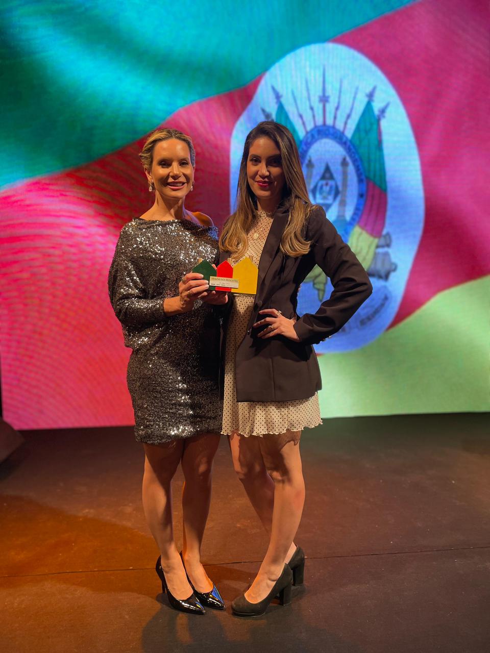 Duas mulheres seguram juntas o troféu da homenagem Heróis das Enchentes, no palco do evento, diante de um painel com a bandeira do Rio Grande do Sul