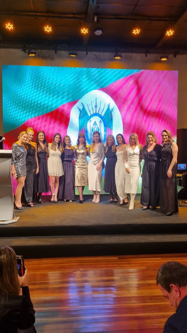 Mulheres lado a lado no palco durante a homenagem, diante do painel com a bandeira do Rio Grande do Sul